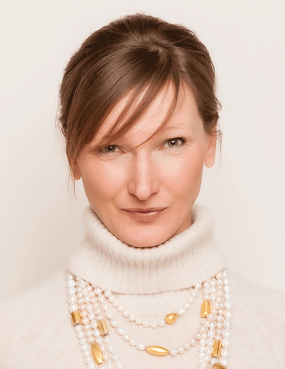 Professionelles Porträt eines Coaches mit warmem Lächeln und selbstbewusstem Blick. In einem cremefarbenen Rollkragenpullover und eleganter Perlenkette vermittelt das Bild Vertrauen, Kompetenz und Stil. Ihr Blick und das warme Licht verleihen dem Bild eine edle und stilvolle Ausstrahlung.